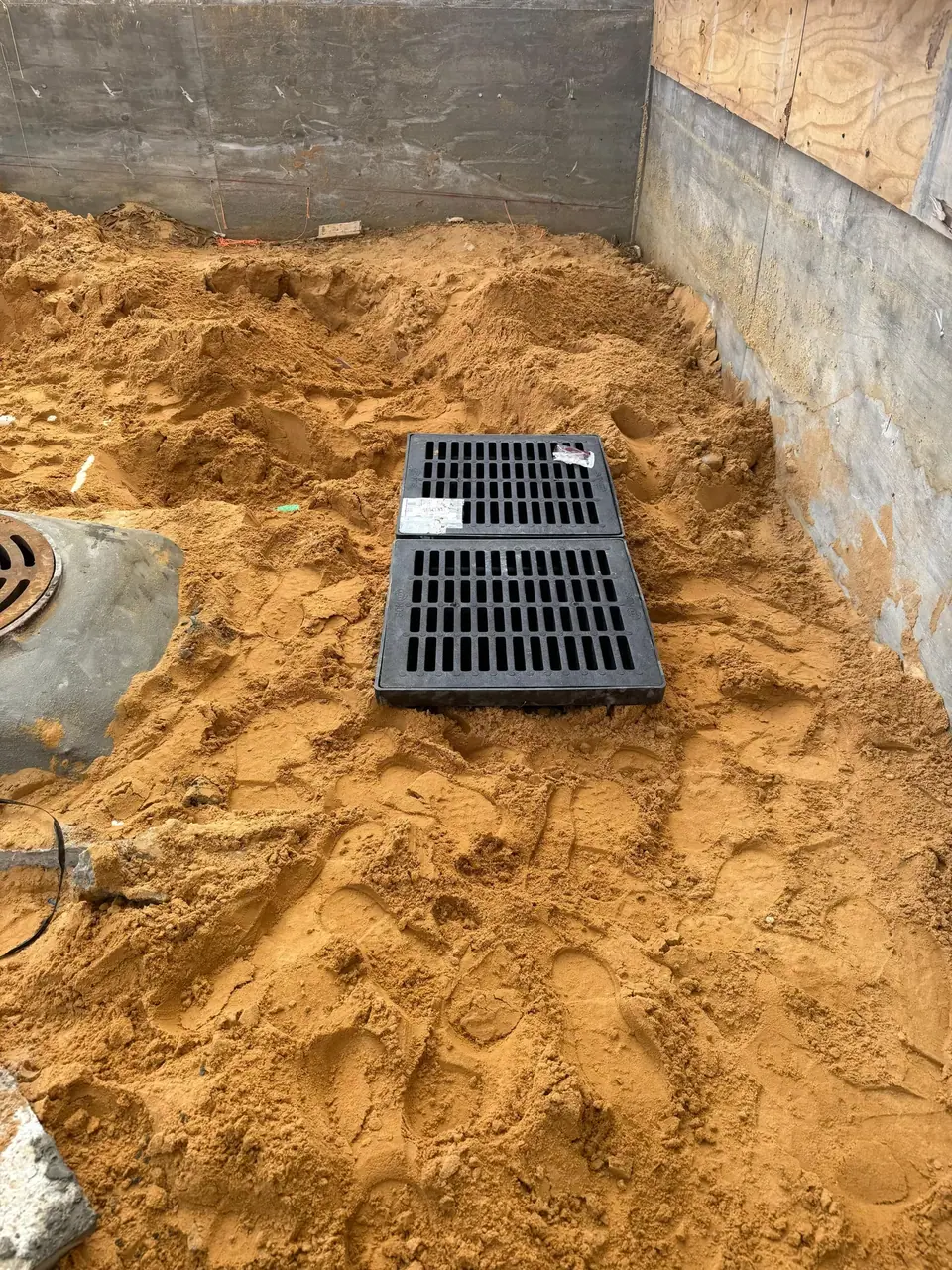 A construction site floor covered in a large pile of sand, featuring two black rectangular grates and a round metal manhole cover. Concrete walls and some plywood are visible in the background.