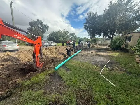 An orange Kubota excavator digs a trench in a grassy area, with a long teal pipe lying nearby. Several workers are present, and parked cars and a mobile home are visible in the background under a cloudy sky.