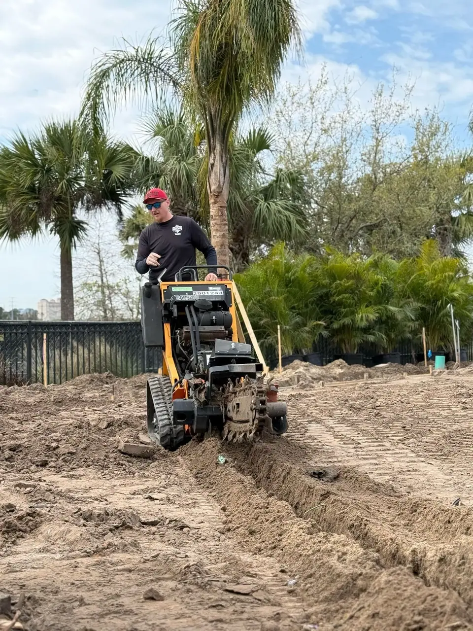 A worker in a red cap and sunglasses operates a yellow trenching machine, digging a furrow in a dirt field. Palm trees and other foliage are visible in the background under a cloudy sky.