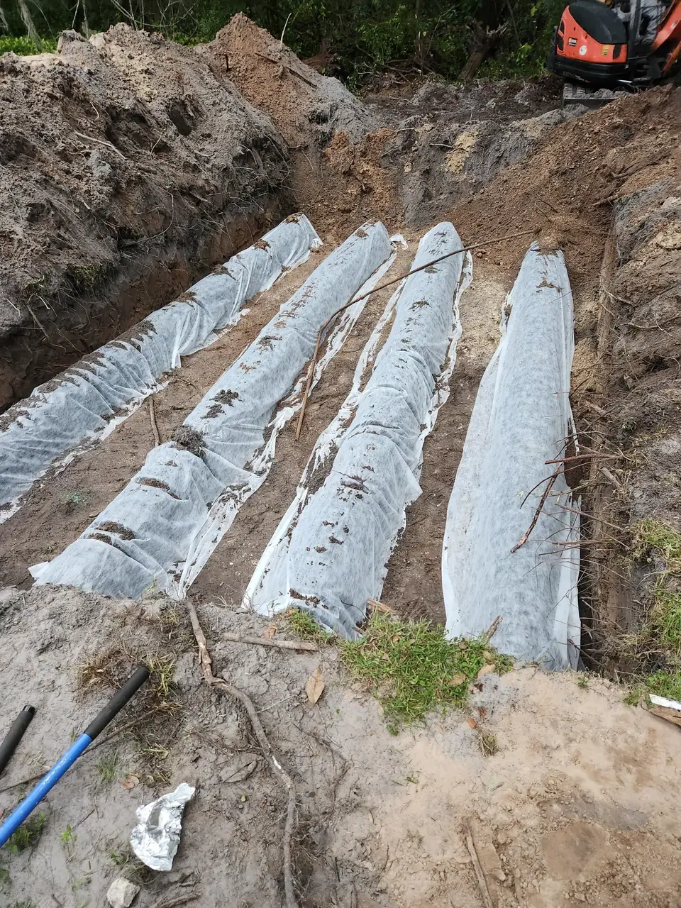 A deep trench in the earth contains several long, parallel objects covered in white fabric, likely part of a drainage or septic system installation. An orange excavator is visible in the background.
