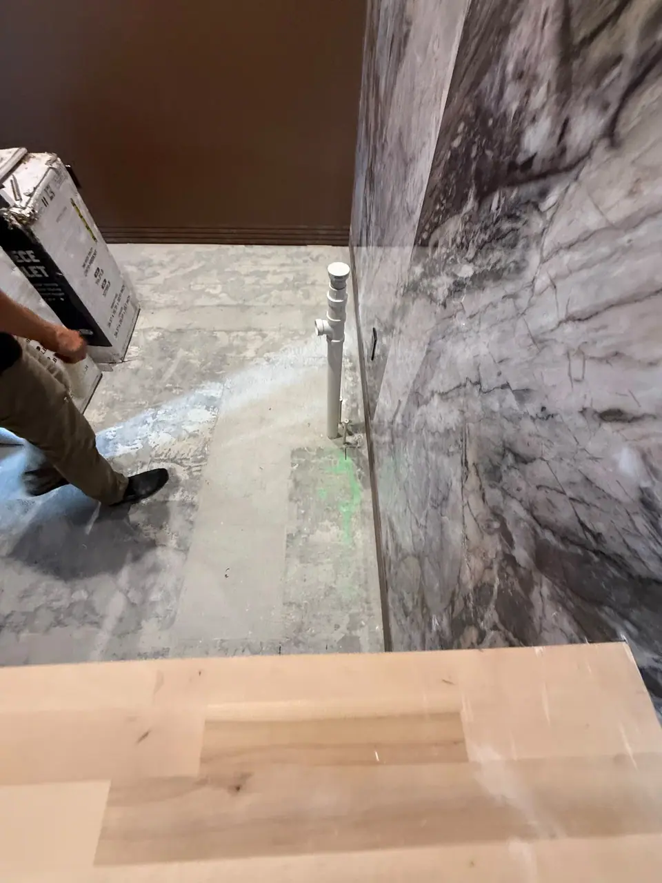 A person in tan pants and dark shoes carries a large white box across a concrete floor. A white pipe extends from the floor against a marbled wall, with a light wooden surface in the foreground.