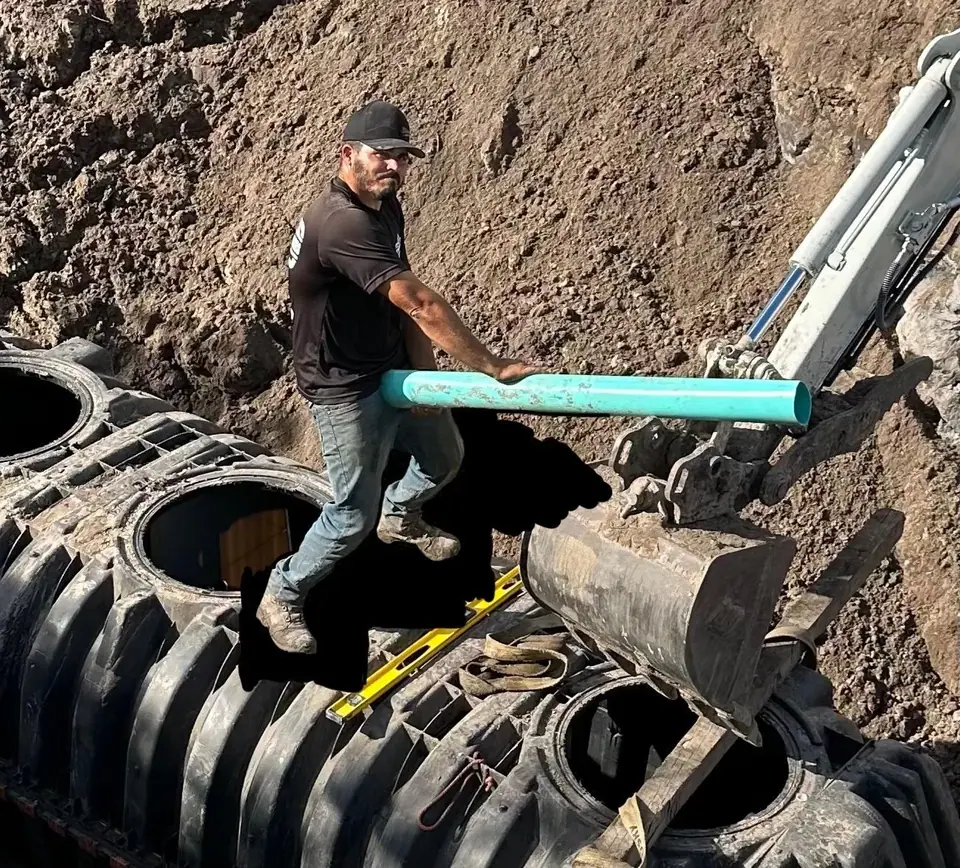 A plumber in a black shirt and cap stands on a large black ribbed tank in an excavated area, holding a light blue pipe. An excavator arm and a dirt wall are visible in the background.