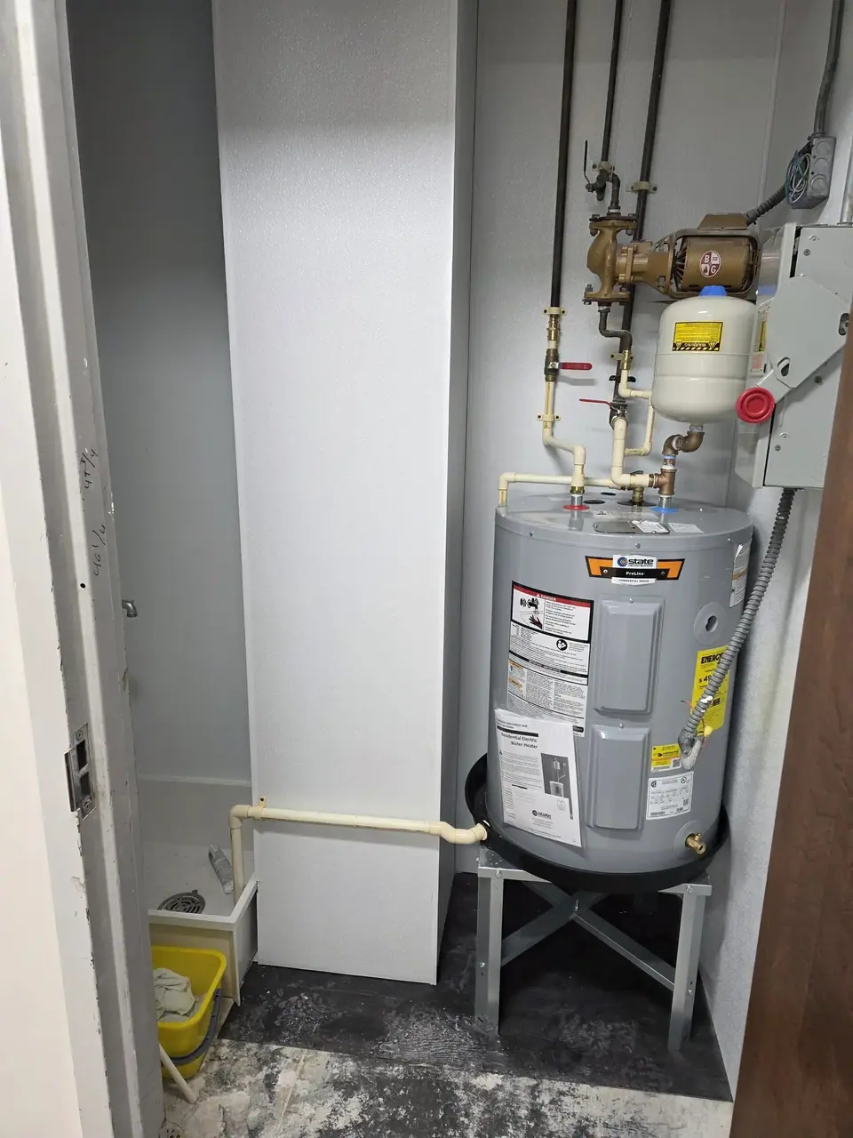 A utility room features a gray electric water heater on a metal stand, connected to an array of pipes, valves, an expansion tank, and a pump. To the left, a small utility sink with a yellow bucket is visible next to a white vertical column.