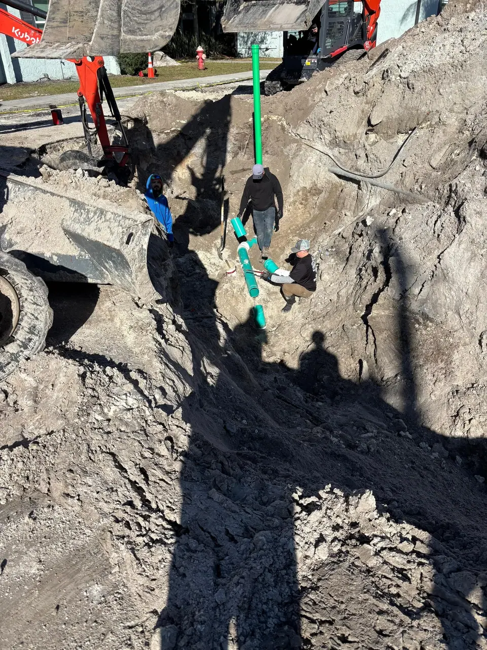 Three construction workers are in a deep trench, installing green pipes. An excavator is partially visible on the left, with piles of dirt surrounding the work area.
