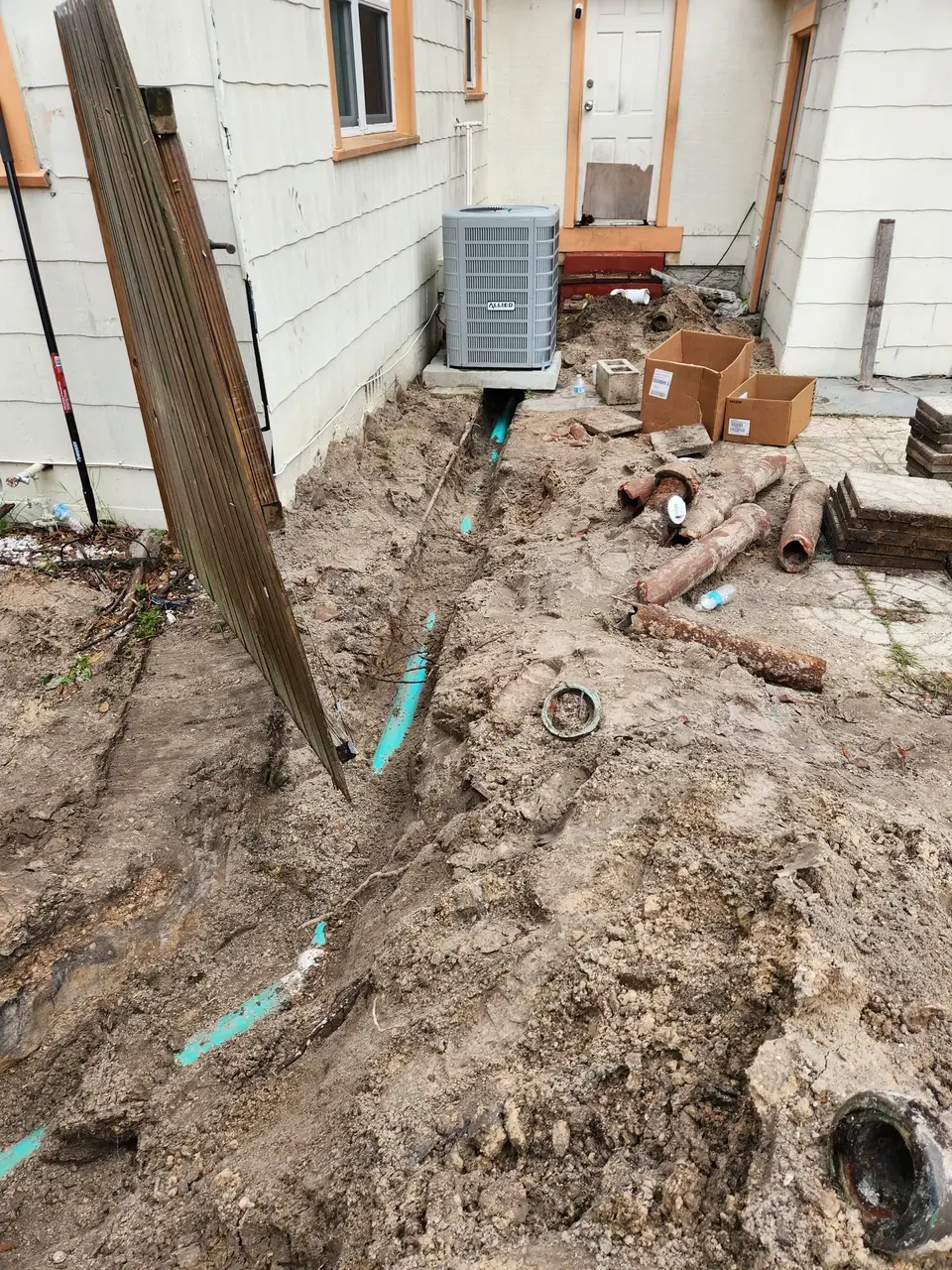 A trench is dug in the dirt next to a house, revealing new blue pipes. Old, rusty pipes, cardboard boxes, and an AC unit are visible around the excavation.