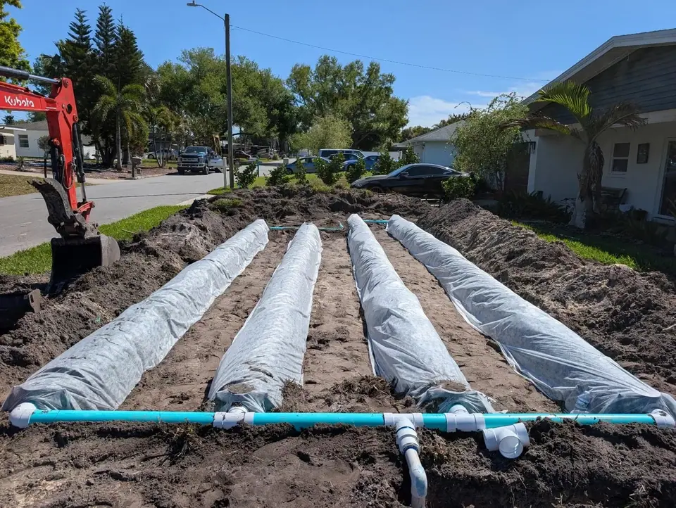 An outdoor photo shows four long, fabric-covered drainage pipes laid in a trench, connected by a blue PVC pipe in the foreground. A red Kubota excavator is visible on the left, with a residential street and houses in the background under a clear blue sky.
