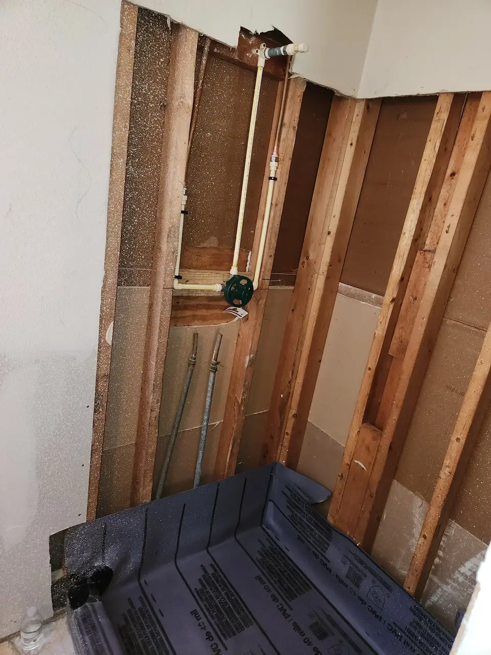 An unfinished bathroom or shower area shows exposed wall studs with plumbing pipes, including a shower valve. A dark gray waterproof membrane is partially installed on the floor, indicating ongoing construction.
