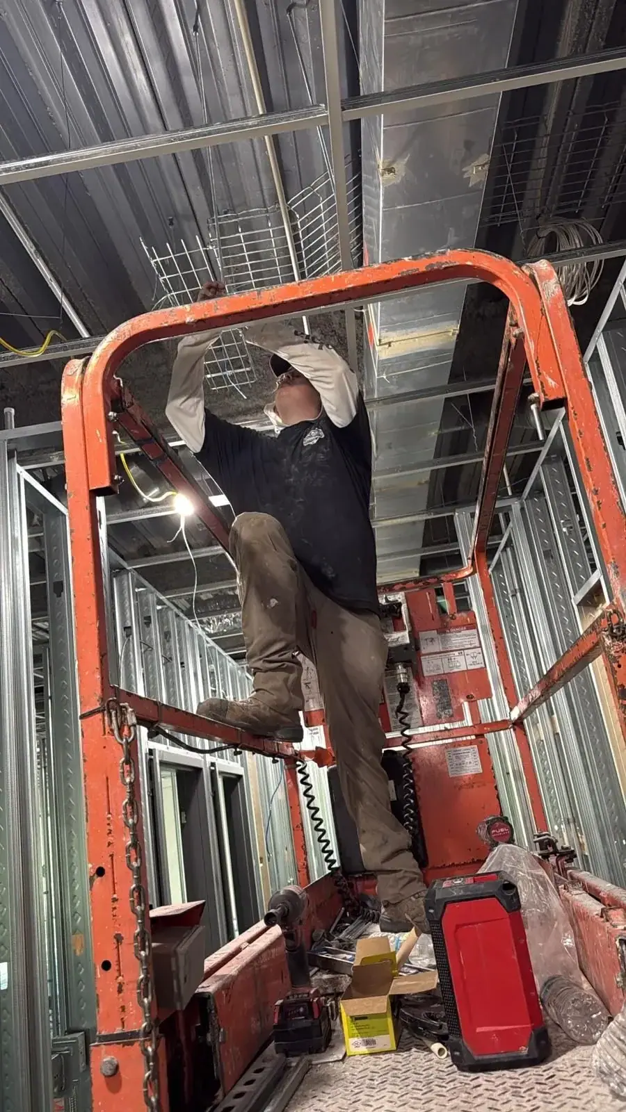 A worker on an orange scissor lift is installing a wire mesh cable tray system on the ceiling of a building under construction. Exposed metal studs and ductwork are visible in the background.