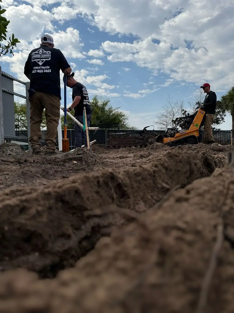 Three men are working outdoors, with two wearing "Plumbing Services" shirts. One man operates a yellow trenching machine while another holds a shovel near a newly dug trench in the foreground.