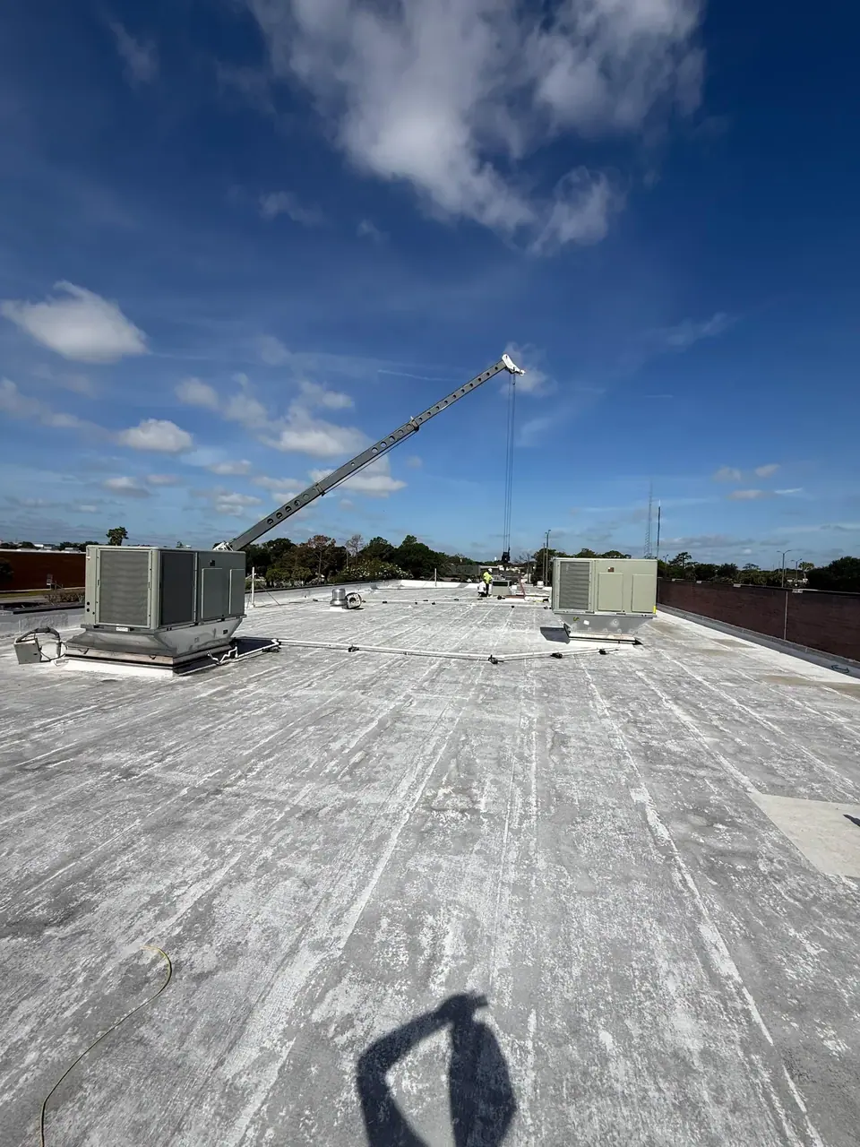 A wide shot of a commercial rooftop with a large crane boom extending over it, positioned to lift or lower HVAC units. Two large, gray HVAC units are visible on the weathered gray roof under a blue sky with scattered clouds.