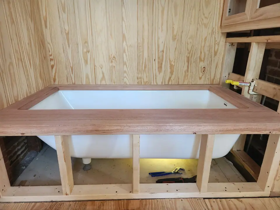 A white bathtub is partially installed within a wooden frame, topped with a finished wooden trim. The walls behind it are covered in vertical wooden paneling, and plumbing and tools are visible around the unfinished installation.