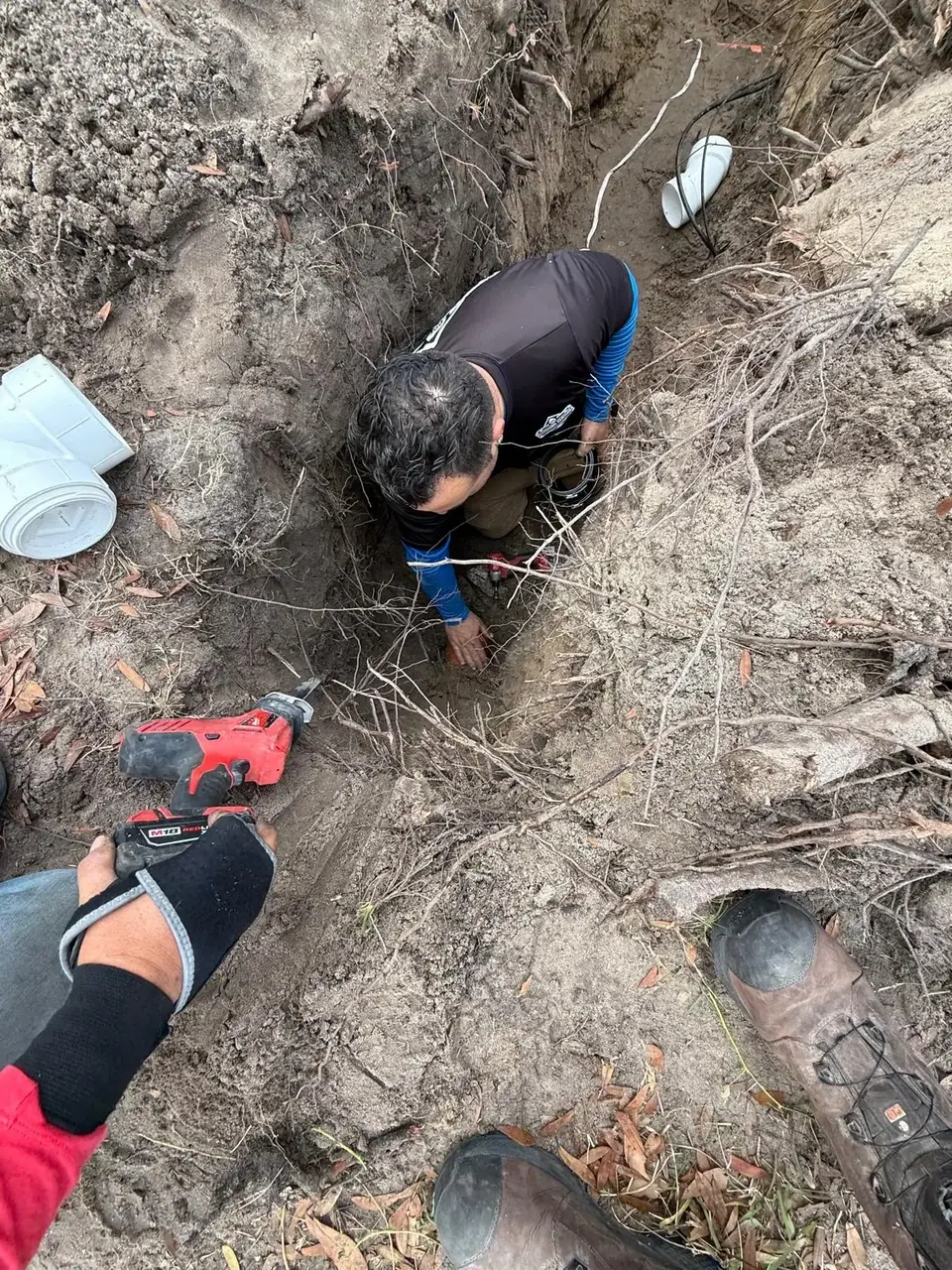 An overhead view shows a plumber in a black and blue shirt working in a dirt trench, with another person's hand holding a red reciprocating saw in the foreground. Plumbing pipes and tools are visible around the excavation.