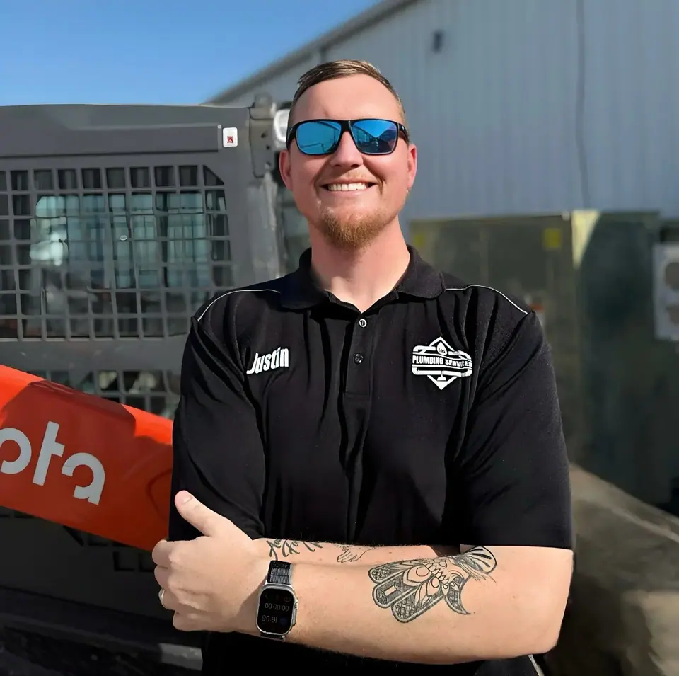 A smiling man wearing sunglasses and a black polo shirt with 'Justin' and a 'Plumbing Services' logo stands with his arms crossed in front of a Kubota skid steer loader. He has tattoos on his forearms and is wearing a smartwatch.