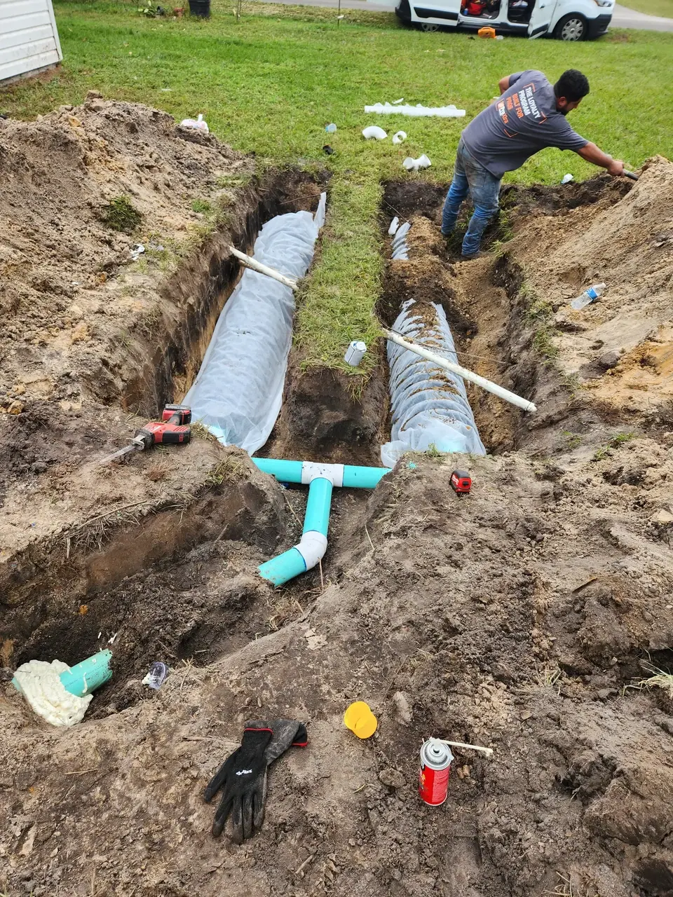 A man works in a trench in a grassy yard, where two long, white-wrapped corrugated pipes are laid for a drainage or septic system. Teal PVC pipes and various tools are scattered around the dug-up area.