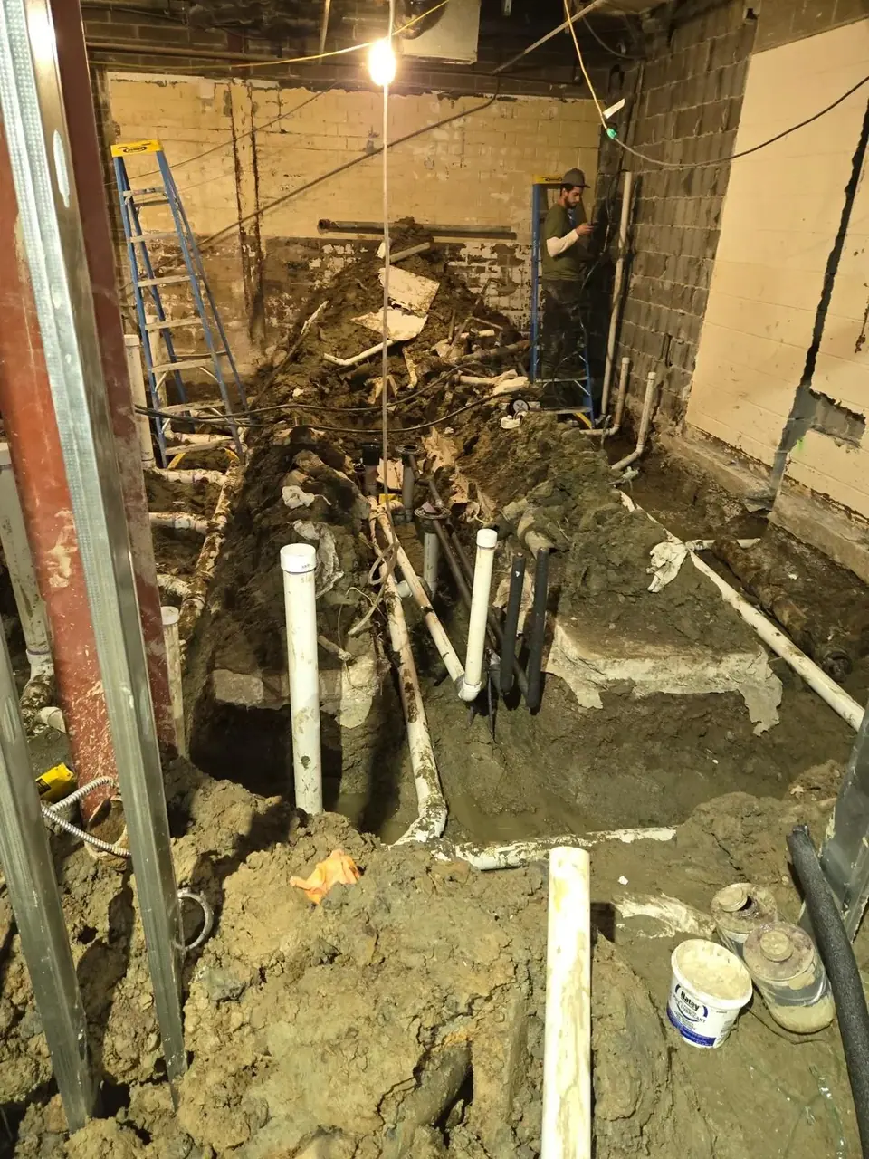 A wide shot of a plumbing job site shows a deep trench dug into a dirt floor, revealing numerous white PVC and black pipes. A worker on a blue ladder is visible in the background, attending to pipes against a cinder block wall, with piles of excavated dirt surrounding the work area.