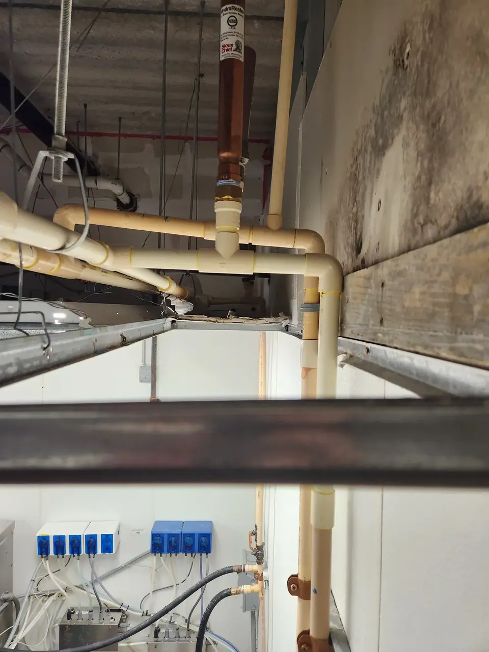 An overhead view of a utility space showing a network of beige and copper pipes, an exposed concrete ceiling, and a wall with dark stains. Below, several blue and white dosing pumps are mounted on a white wall, connected by various tubes and wires.