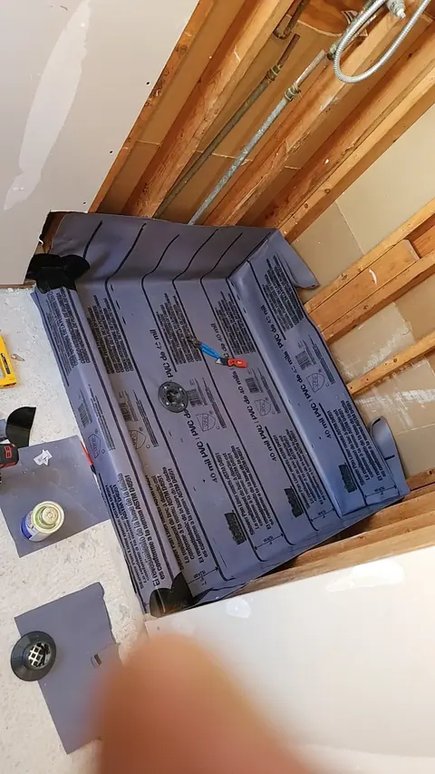A gray PVC shower pan liner is installed in a partially constructed shower area, with exposed wooden studs and pipes visible above. Various tools, including pliers, a drill, and an adhesive can, are on the floor around the liner, next to a shower drain component.