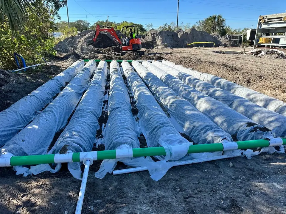 Multiple large, white-wrapped pipes are laid in a trench at a construction site, with a green and white pipe assembly visible in the foreground. An excavator is actively digging in the background, surrounded by piles of dirt.