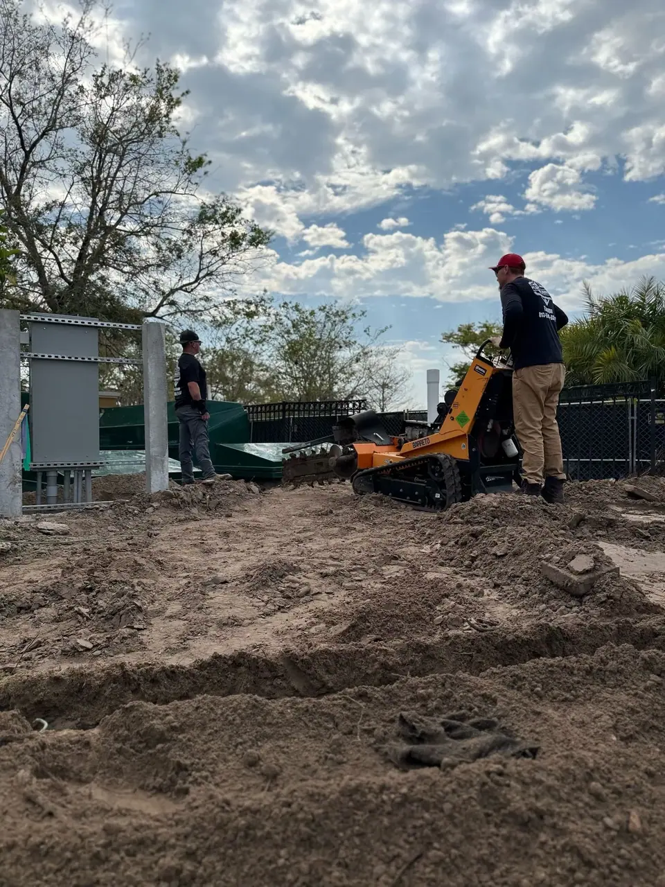 Two men are working on a construction site under a cloudy sky. One man operates an orange stump grinder on a mound of dirt, while another stands near green structures and metal panels in the background.