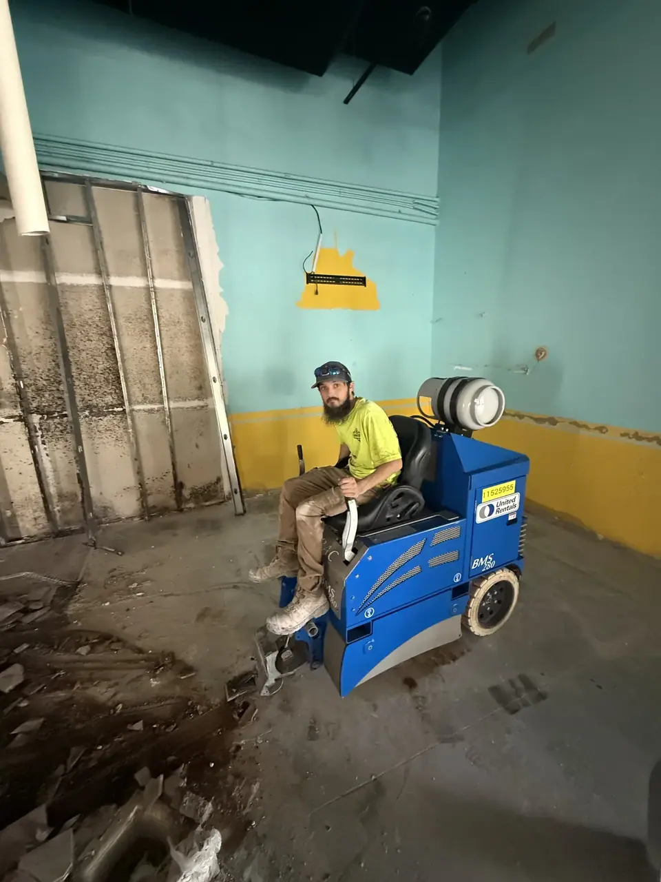 A man with a beard and baseball cap sits on a blue ride-on floor scraper machine, identified as a United Rentals BMS 280, in a room under renovation. The room features light blue and yellow walls, exposed metal wall framing, and a concrete floor with debris.
