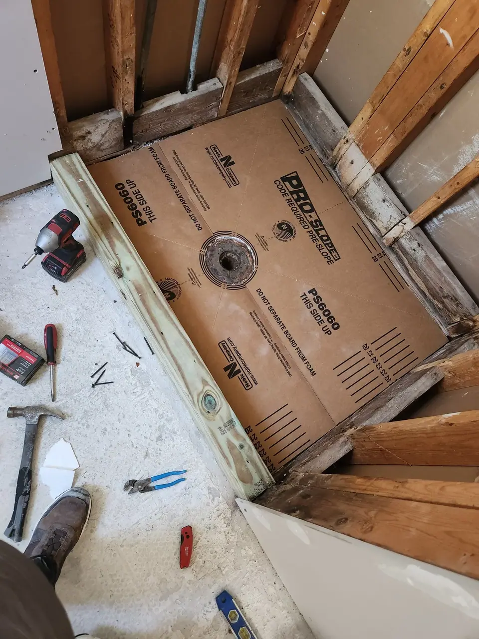 An overhead view of a shower pan installation in progress, showing a brown 'PRO-SLOPE PS6060' pre-sloped shower base with a central drain hole, surrounded by wooden framing. Various construction tools like a drill, hammer, and nails are scattered on the concrete floor around the installation.