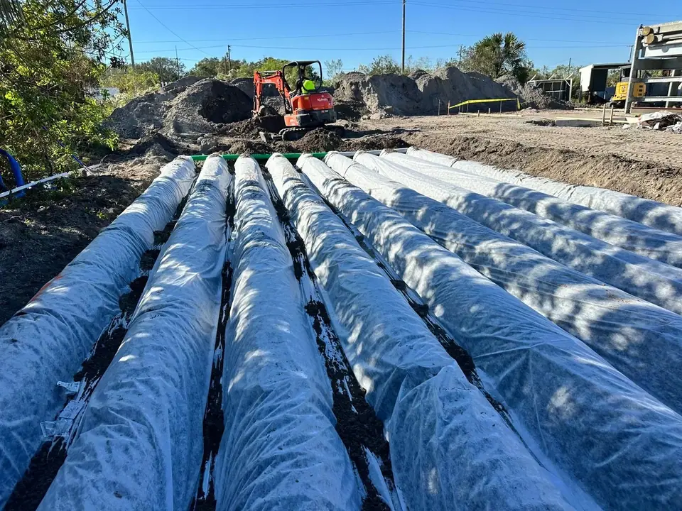 Multiple large pipes, covered in white fabric, are laid in trenches at a construction site. An orange excavator is visible in the background, surrounded by piles of dirt under a clear blue sky.