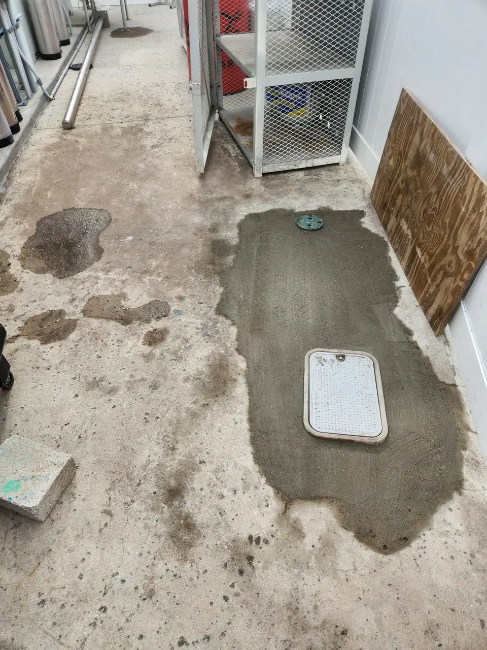 A concrete floor with several wet spots and a fresh patch of darker gray concrete containing a round green cover and a rectangular white utility cover. Industrial pipes, a metal mesh cage, and a piece of plywood are visible in the background.