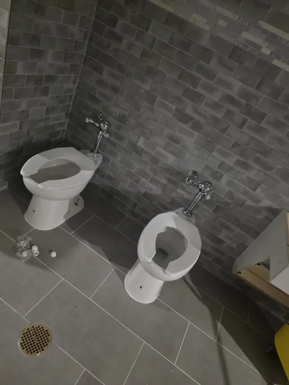 Two white toilets with open seats are positioned on a light gray tiled floor against dark gray subway tile walls. Scattered white plastic caps are visible on the floor near the left toilet, and a floor drain is in the bottom left.