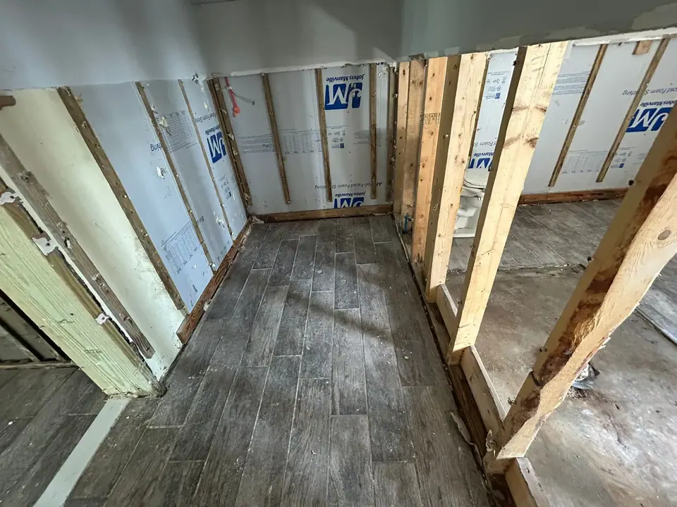 An interior view of a room under renovation, featuring exposed wooden wall studs, white insulation panels with 'Johns Manville' branding, and dark grey wood-look floor tiles. A toilet base is partially visible behind some studs on the right.