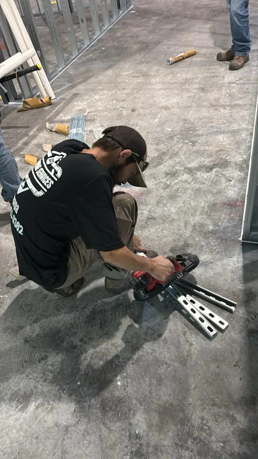 A worker wearing a black t-shirt and a baseball cap crouches on a concrete floor, using a red power tool to cut metal channels. Metal framing studs are visible in the background of the construction site.