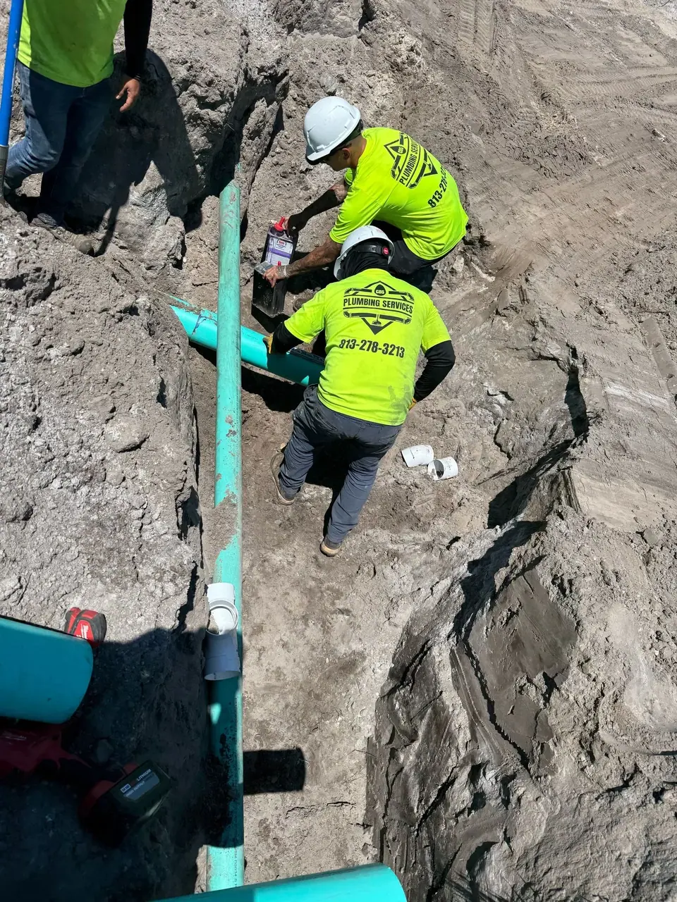 Two plumbers in hard hats and neon yellow shirts are working in a dirt trench, installing or repairing turquoise pipes. One worker is applying a substance from a purple container to a pipe section.