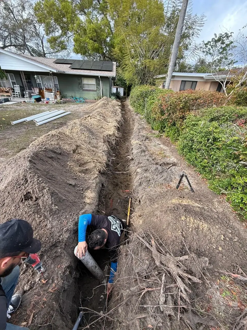 Two plumbers are working in a long, deep trench in a residential yard. One plumber is in the trench, handling a large pipe, while the other kneels at the edge with a power tool, with excavated dirt piled alongside the trench.
