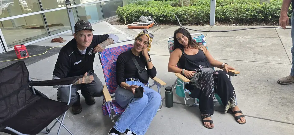 Three people are gathered outdoors on a concrete surface. A man crouches on the left, while two women sit in beach chairs, all smiling towards the camera, with construction equipment visible in the background.