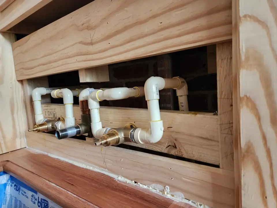 Plumbing pipes, including light-colored PVC and brass fittings, are installed within an unfinished wooden frame. A brick wall is visible behind the pipes, and a blue plastic sheet is at the bottom left.