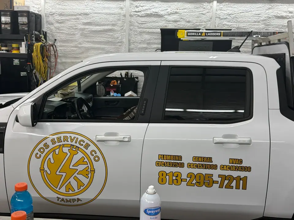 A white service truck for 'CDS SERVICE CO TAMPA' is parked indoors, displaying a logo with a lightning bolt, flame, and snowflake, along with services like plumbing, general, and HVAC, and a phone number. A Gorilla Ladders rack is visible on the roof.