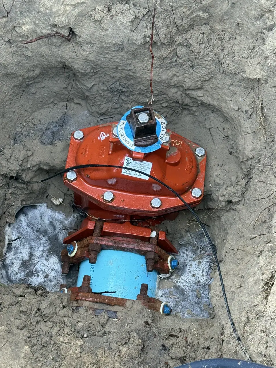 A partially buried orange-red fire hydrant valve assembly is shown in a pit of light-colored soil. It features a blue pipe section at the bottom, connected by rusty bolts, with a black wire and a thin copper wire attached to the top of the valve.