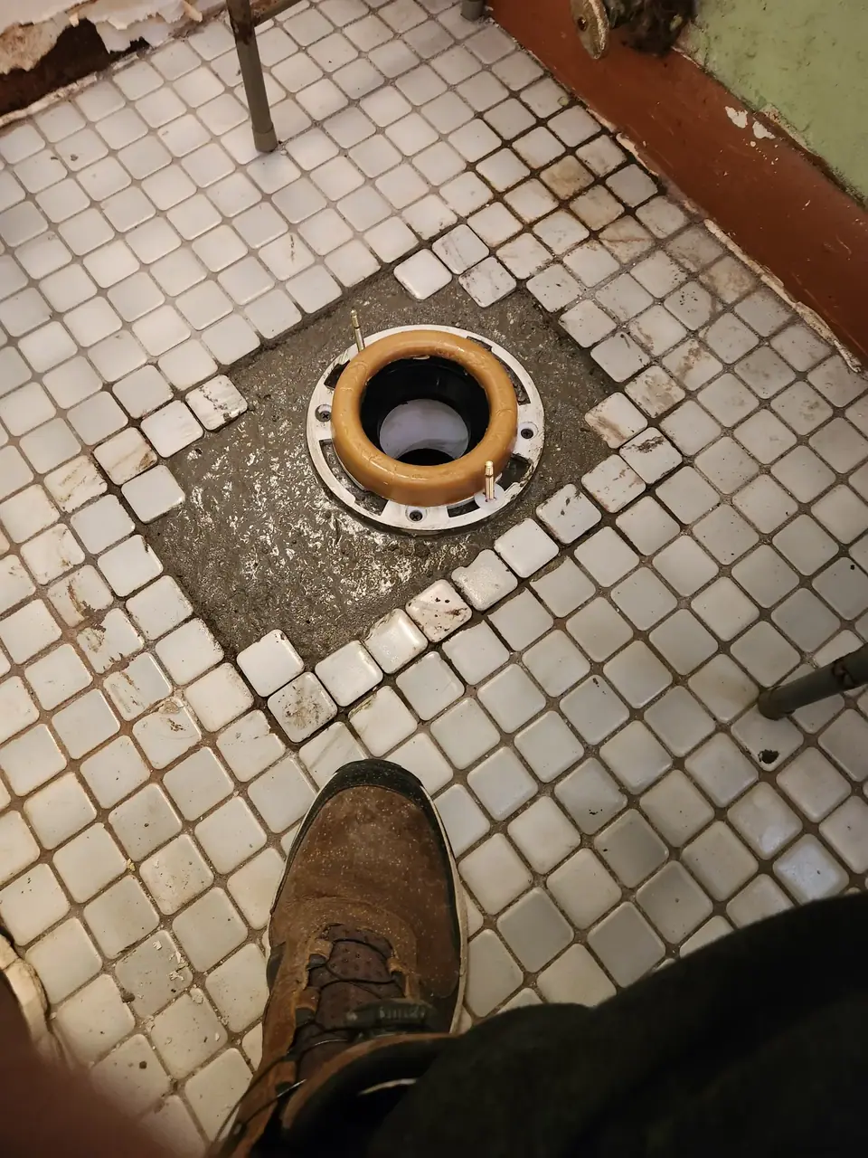 A toilet flange with a wax ring is installed in a bathroom floor, surrounded by missing white square tiles that expose the subfloor. A person's brown shoe and leg are visible in the foreground, looking down at the installation.