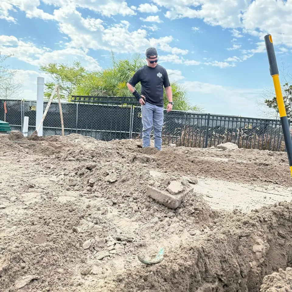 A worker wearing a black shirt, grey pants, cap, and sunglasses stands on a pile of excavated dirt, looking down into a trench. In the background, there is a black fence, green trees, and a blue sky with clouds. A white PVC pipe is visible on the left, and a yellow and black tool is partially visible on the right.
