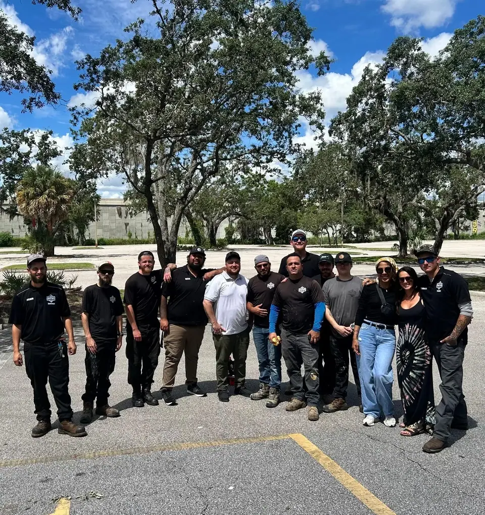 A group of 14 people, mostly men, stand in a paved outdoor area under a blue sky with white clouds. Large trees with Spanish moss are visible in the background.