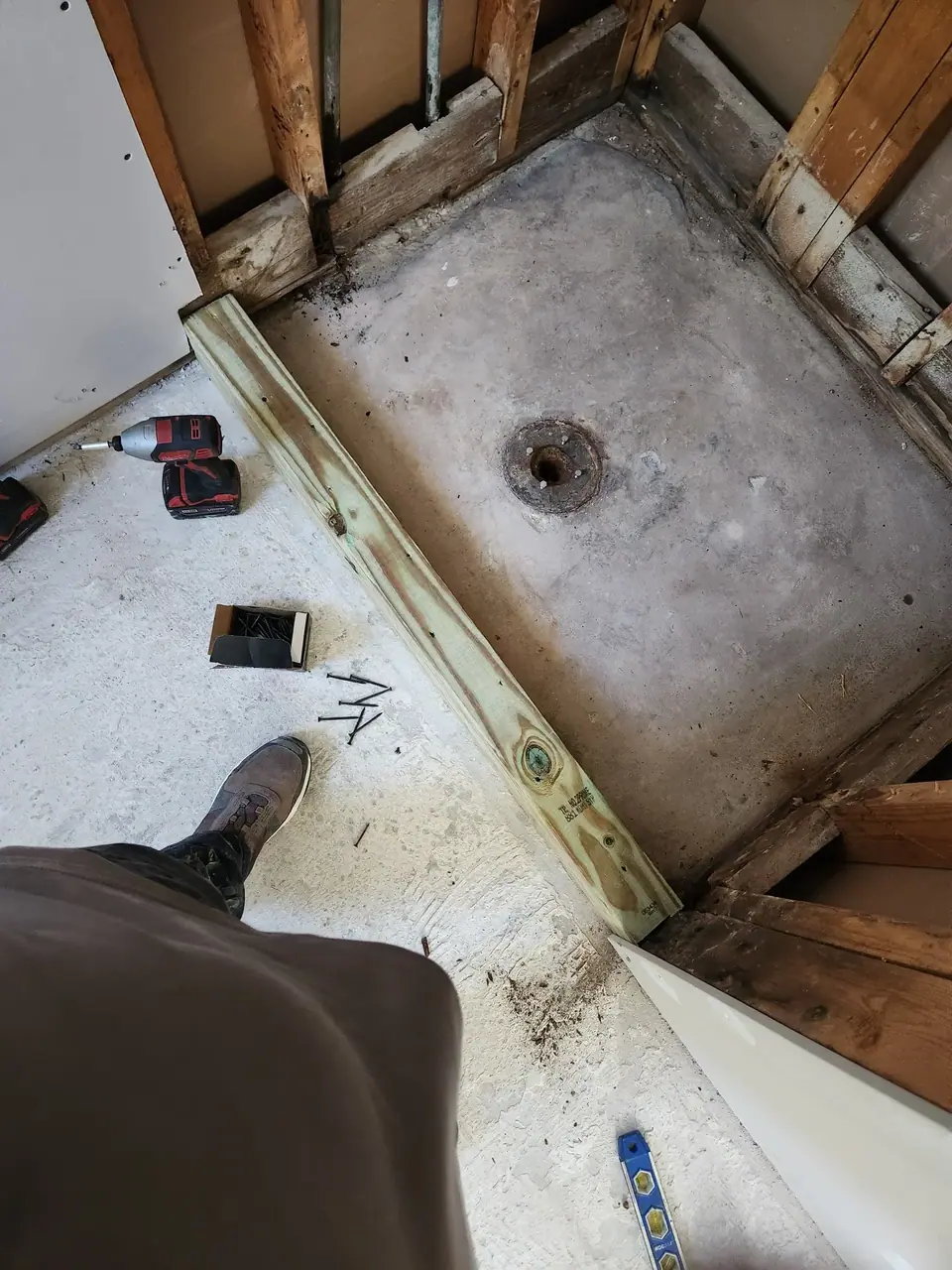 An overhead view of a shower pan under construction, showing a concrete base with a drain, exposed wooden framing, and a treated lumber board laid across the floor. A power drill, screws, and a person's foot are also visible on the concrete.