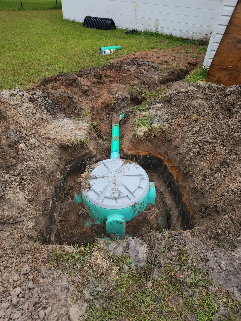 A teal-colored tank with a gray lid is partially buried in a deep trench in a grassy yard. A green pipe extends from the tank, and a white building is visible in the background.