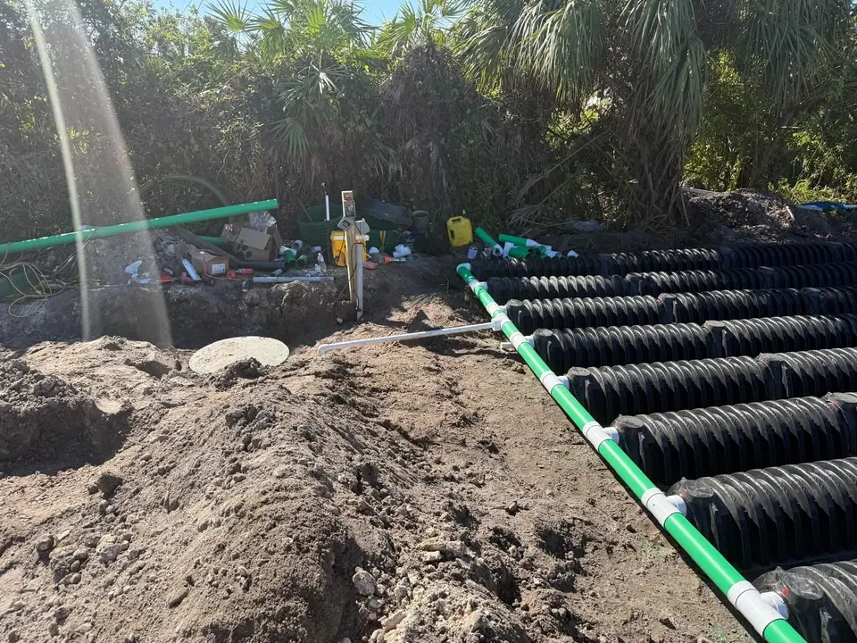 An outdoor plumbing installation featuring green and white pipes connected to rows of black corrugated drainage pipes laid in disturbed earth. A round concrete lid is visible in the foreground, with tools and materials in the background.