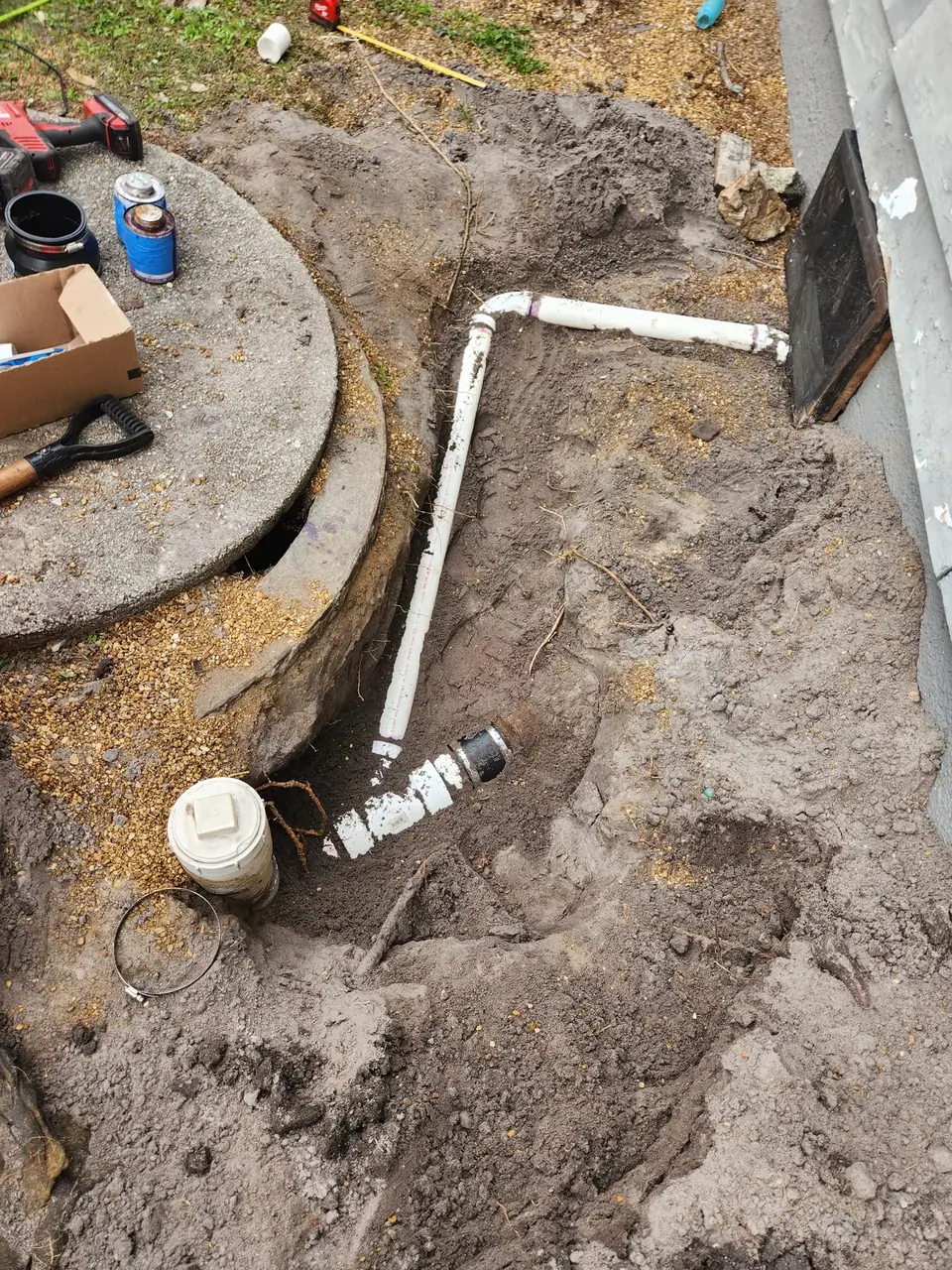 An excavated trench in dirt shows white PVC pipes being installed or repaired, with a concrete manhole cover and plumbing tools nearby.