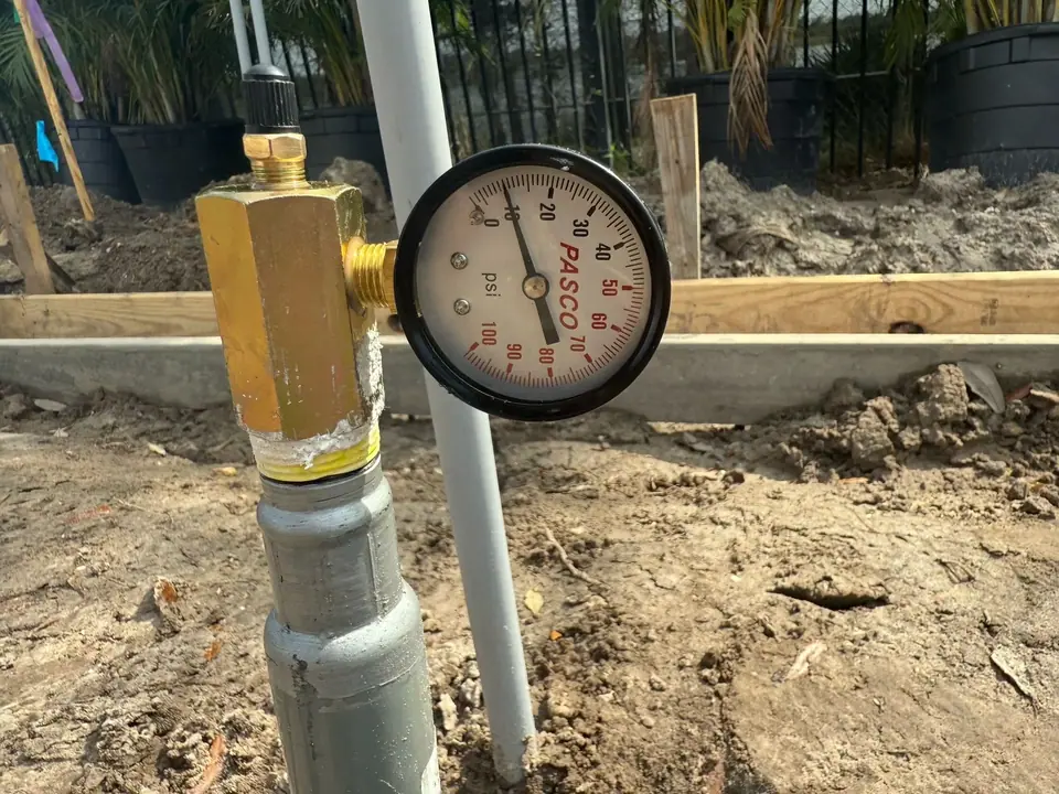 A close-up view of a brass pressure gauge, labeled 'PASCO', attached to a grey pipe partially buried in dirt. The gauge's needle points to approximately 12 psi, with wooden planks and potted plants visible in the background.