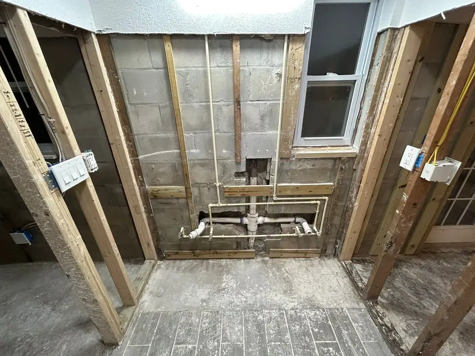 The image displays exposed plumbing pipes, including white PVC and beige PEX, installed against a concrete block wall. Wooden studs frame the area, and a window is visible on the right side, indicating a renovation or construction stage.