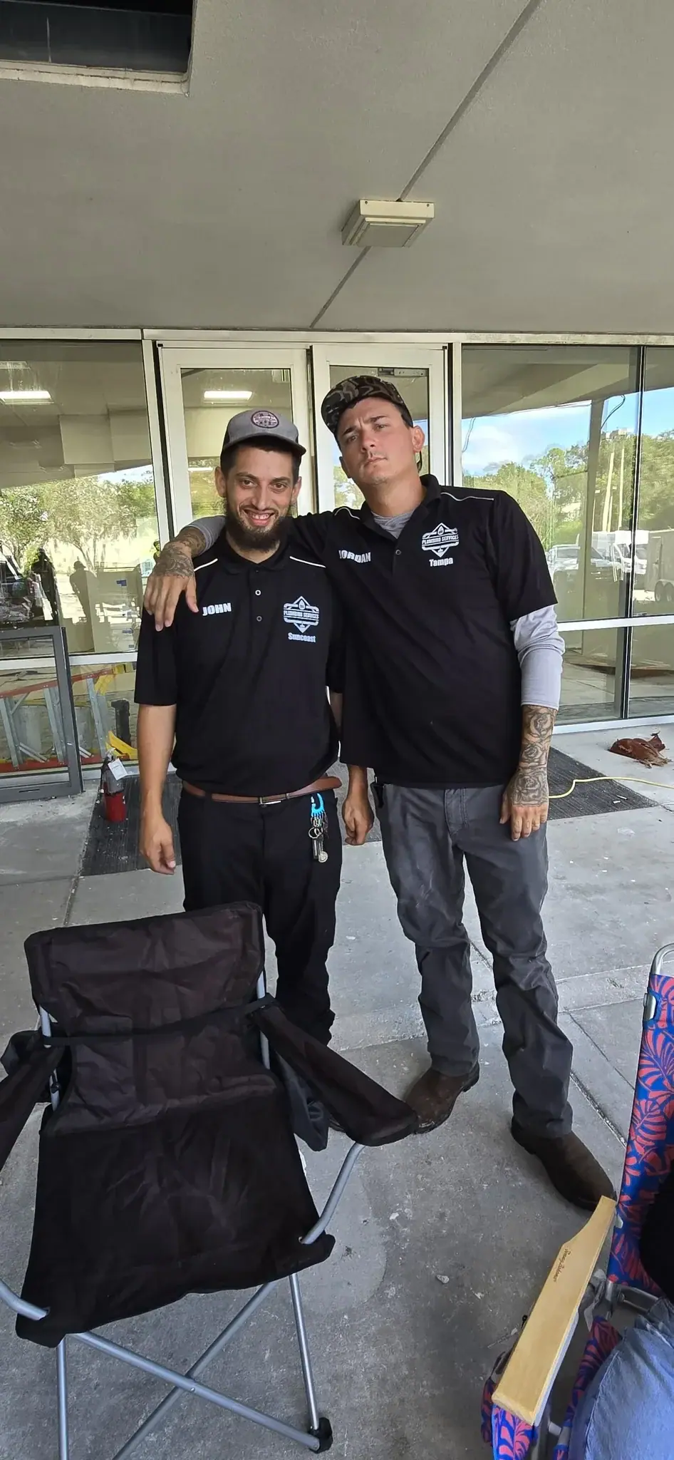 Two men in black polo shirts with company logos stand outside a building with glass doors. The man on the left, with a beard and grey cap, has his arm around the shoulder of the man on the right, who wears a camouflage cap and has tattoos.