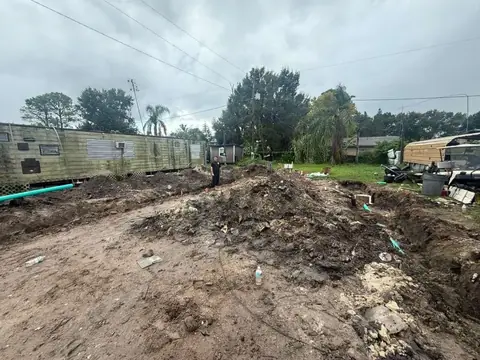A muddy construction site with trenches and dirt piles in the foreground, showing green and white pipes. In the background, a weathered mobile home is on the left, and a partially disassembled RV or trailer is on the right, with two men standing near the trenches under an overcast sky.