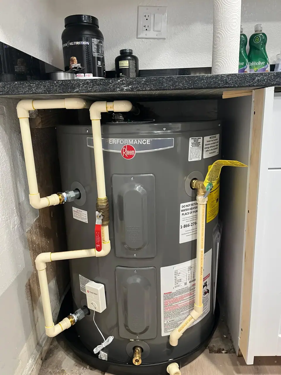 A Rheem Performance water heater is installed under a counter, with CPVC pipes connected to it and a flood alert sensor attached. Various household items, including protein powder and dish soap, are visible on the counter above.
