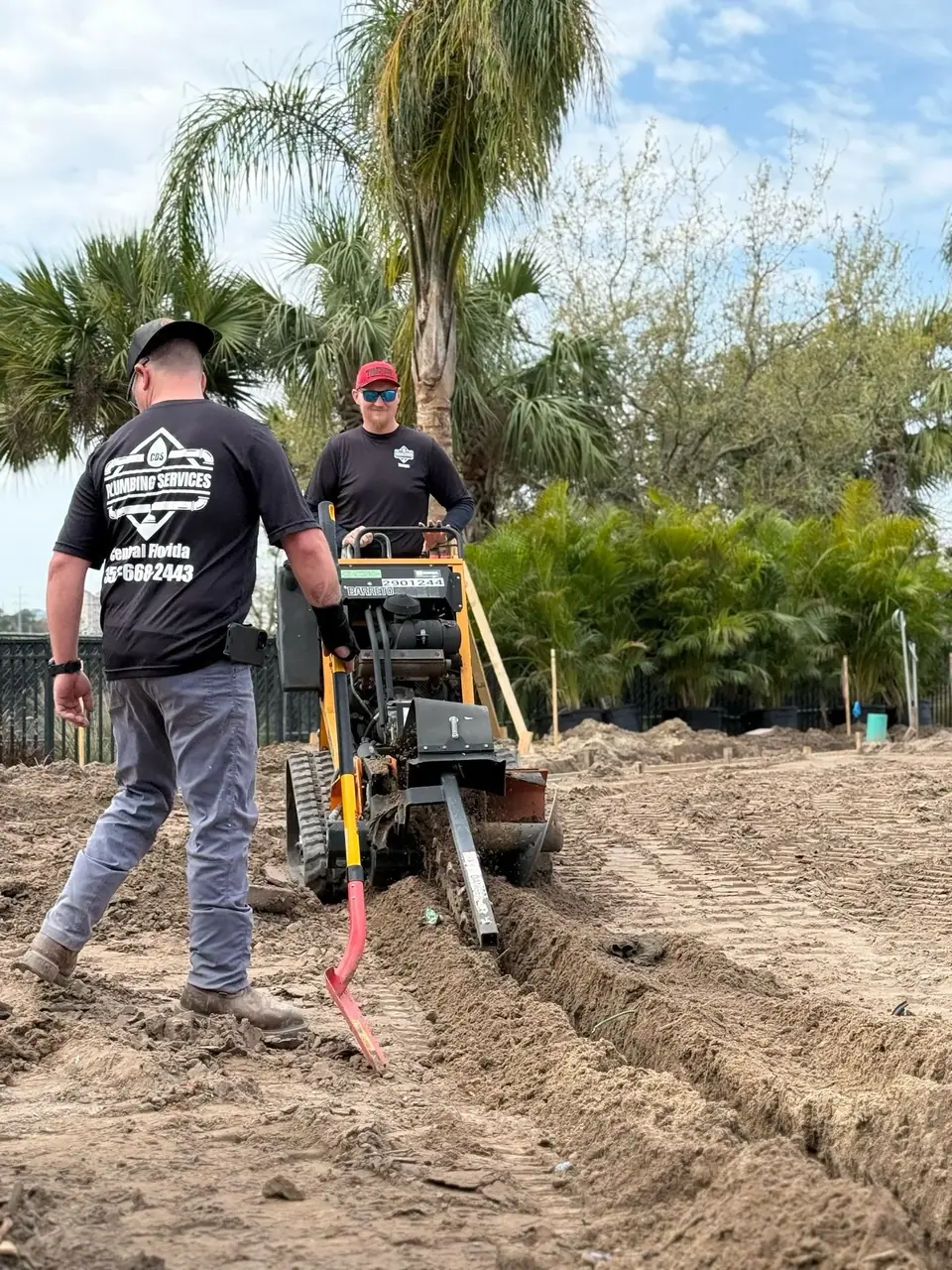 Two workers are shown in a dirt field, one operating a trenching machine to dig a trench, and another worker walking beside it. Palm trees are visible in the background under a partly cloudy sky.