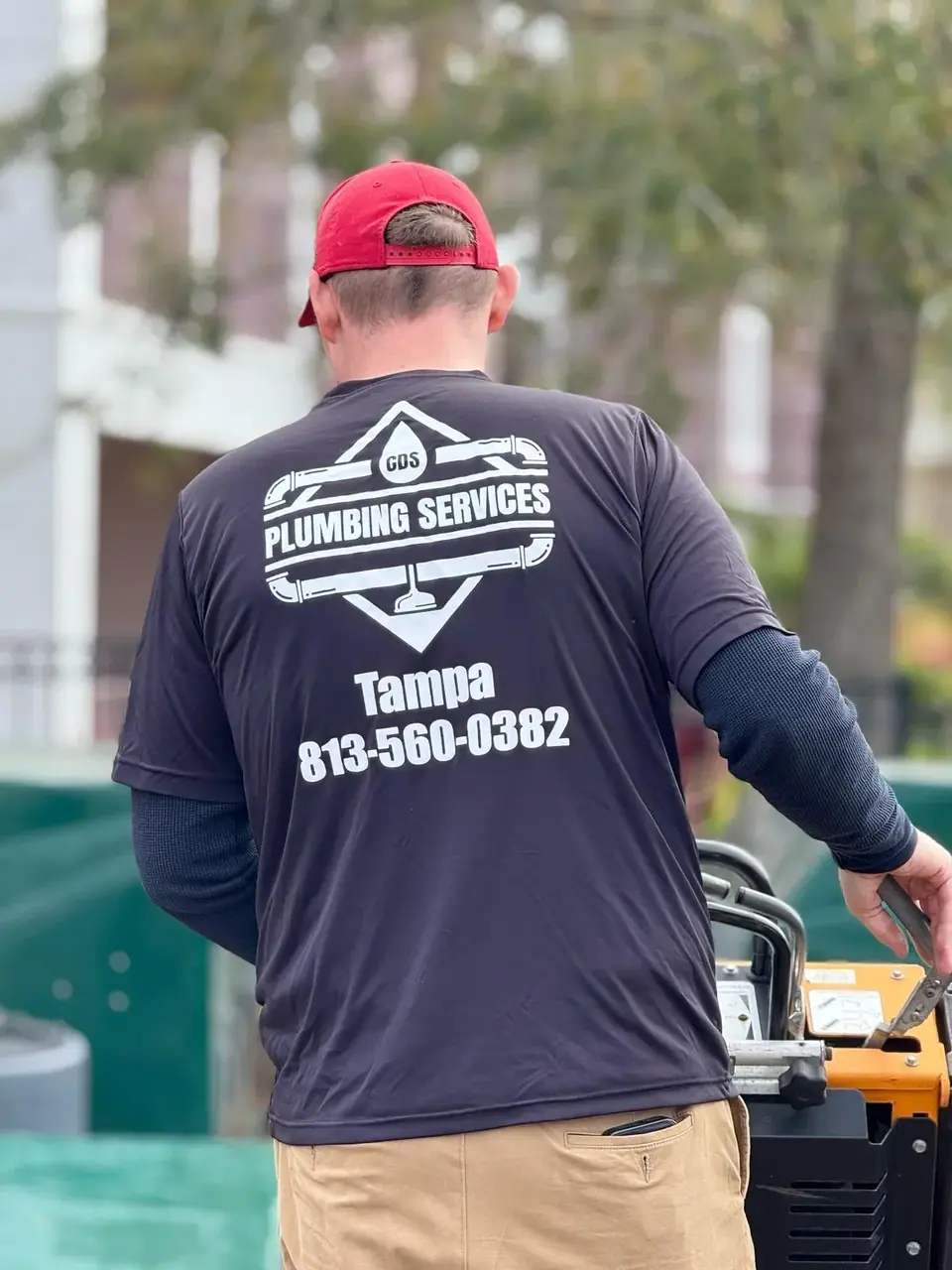 A person wearing a red cap and a dark blue 'CDS Plumbing Services' t-shirt is seen from behind, holding onto orange machinery. The shirt also displays 'Tampa 813-560-0382' and a plumbing logo.